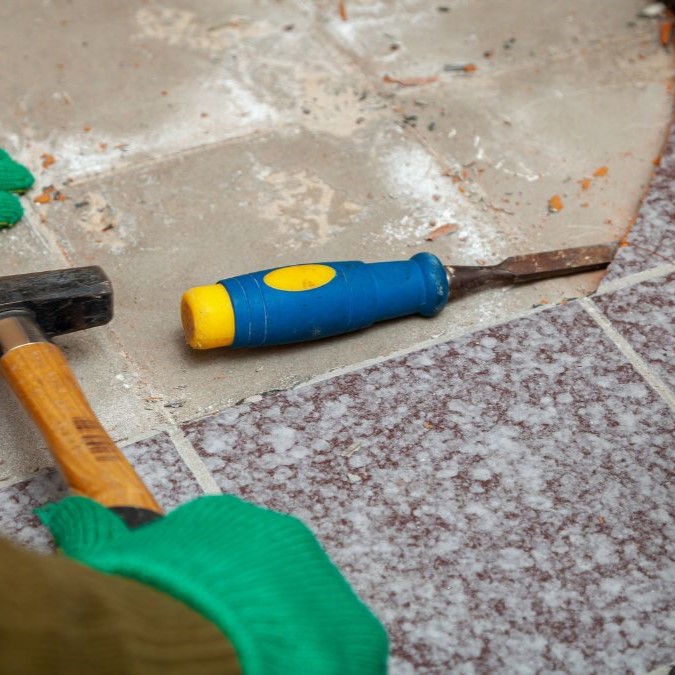 A chisel being hit with a hammer to loosen a tile attached to concrete.